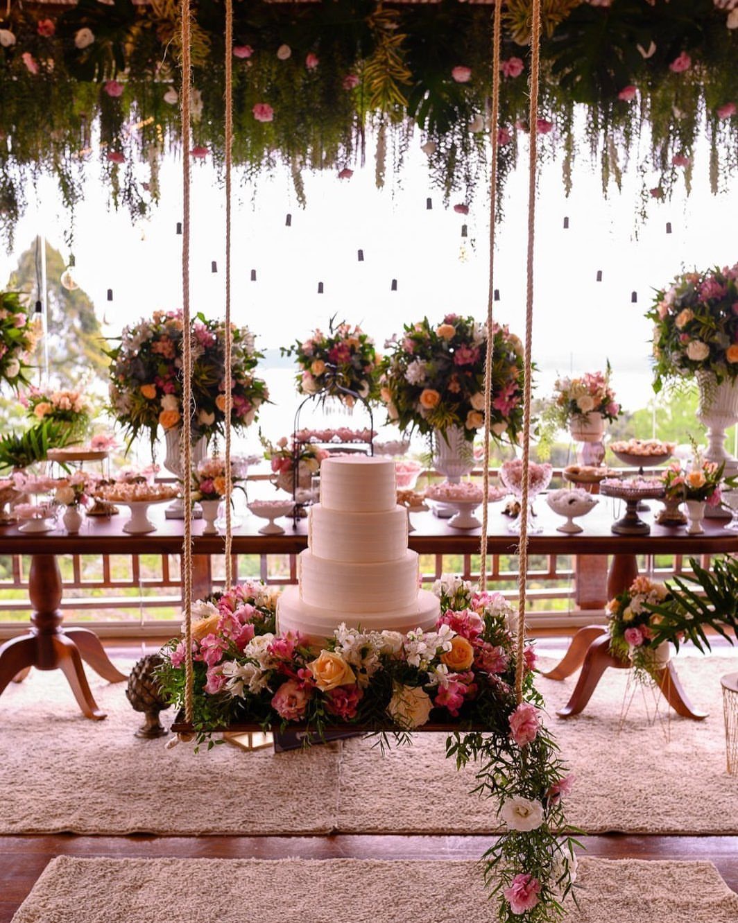 13 Wedding Ideas for Country Ceremonies That Feel Lived-In 4 A white tiered wedding cake displayed on a flower-adorned wooden swing suspended by ropes.