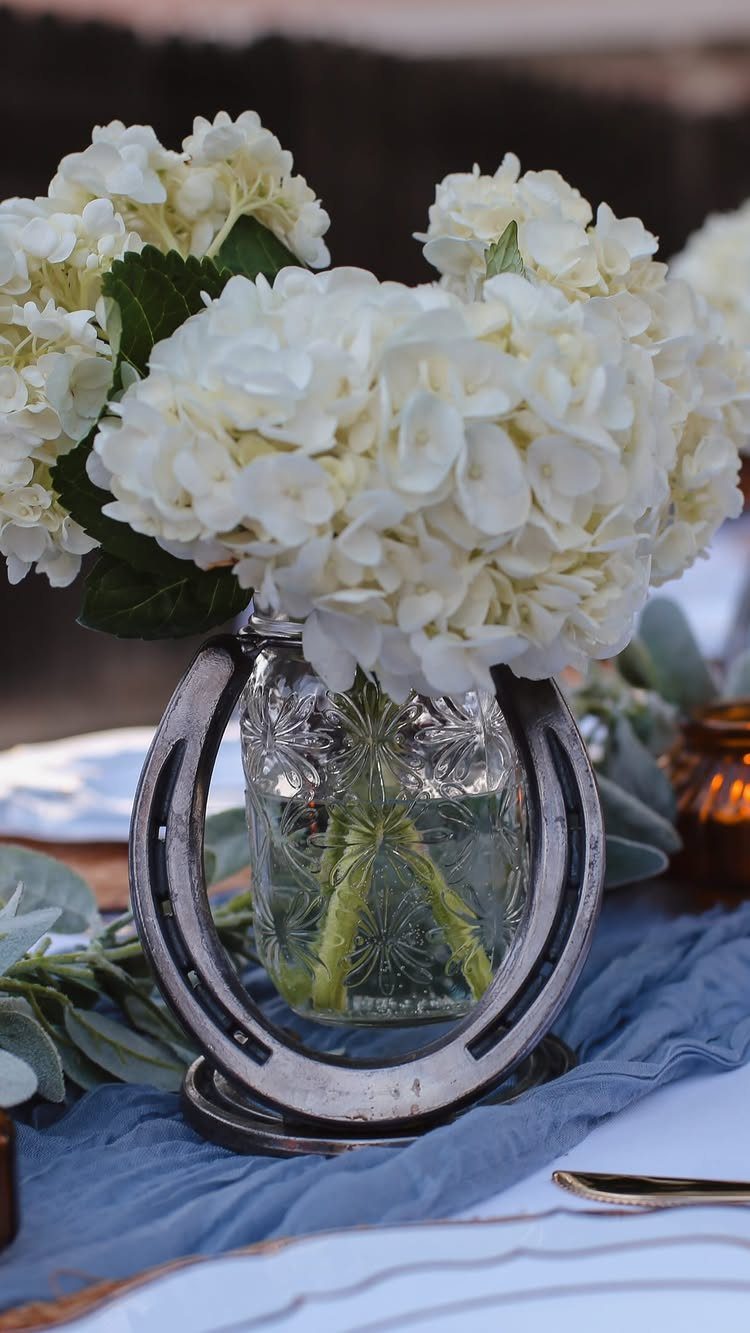 13 Wedding Ideas for Country Ceremonies That Feel Lived-In 11 White hydrangeas in a glass vase framed by a large metal horseshoe on a table.