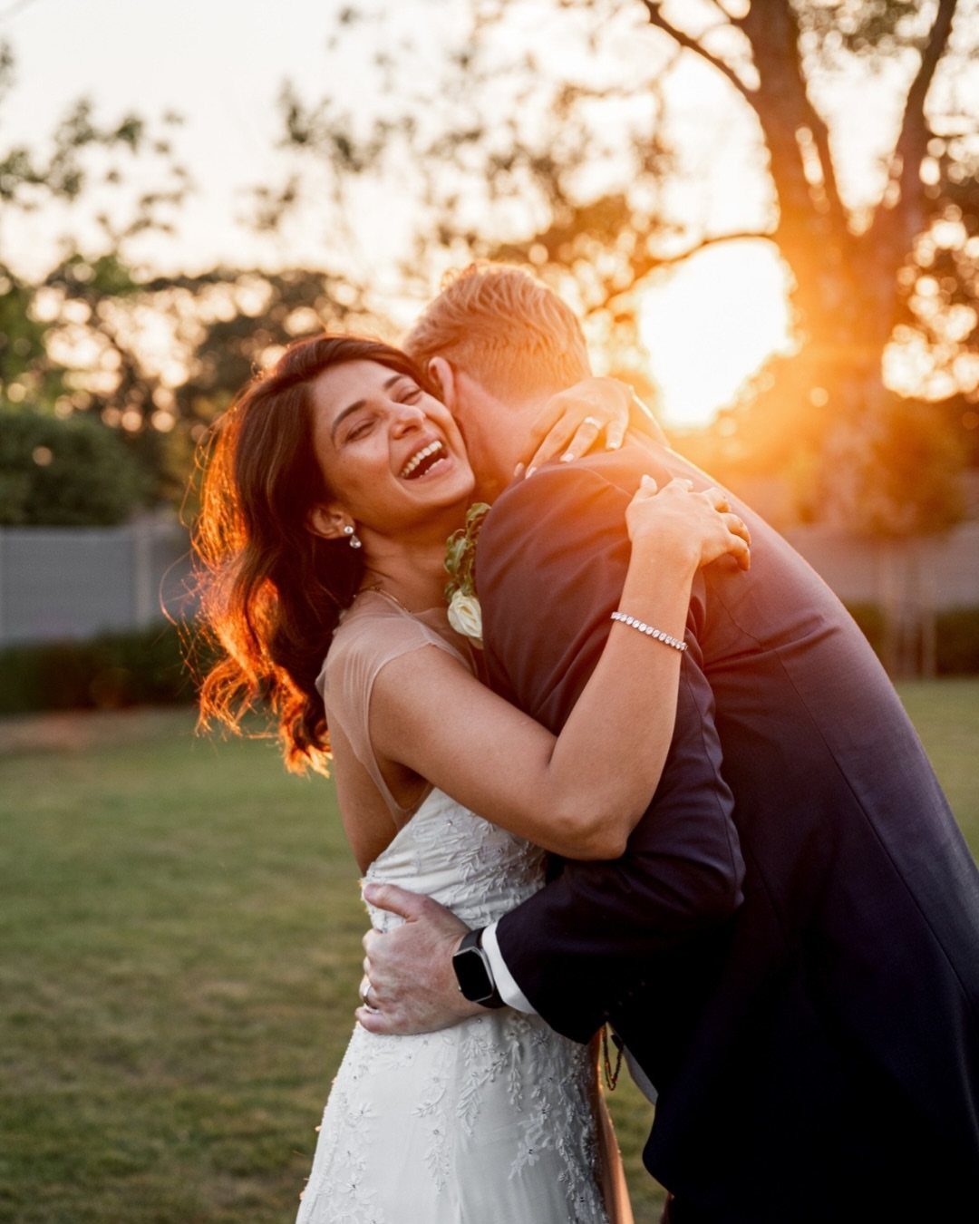 7 Wedding Picture Poses for the First Look, Sunset, and More 5 A laughing bride in a white lace dress embraces her groom in a sunlit grassy field.