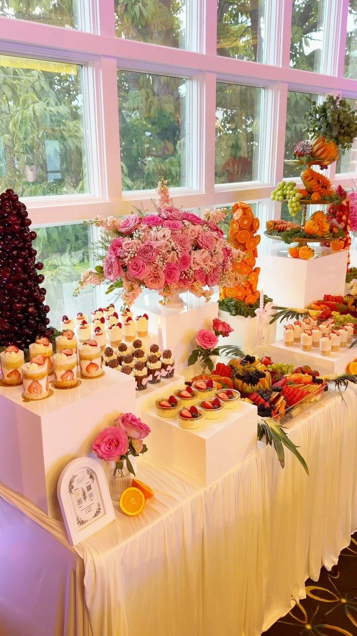 16 Wedding Reception Tables and Layouts for Every Celebration 11 A dessert buffet table with various pastries, fruit displays, and pink flowers in front of windows.
