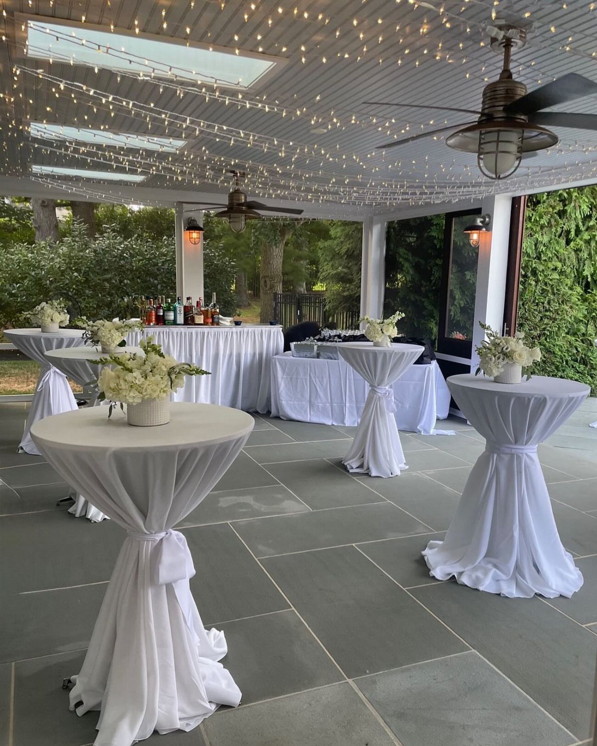 16 Wedding Reception Tables and Layouts for Every Celebration 12 White draped cocktail tables with floral centerpieces arranged on a stone patio under string lights.
