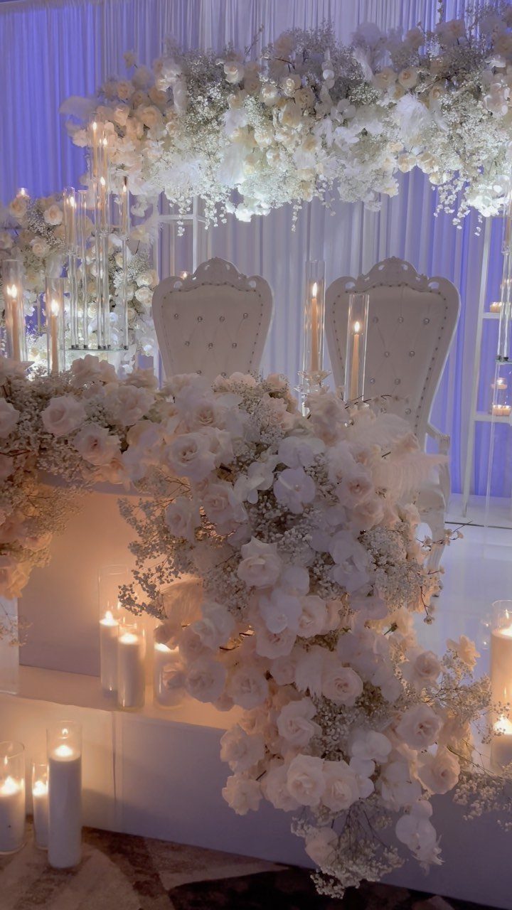16 Wedding Reception Tables and Layouts for Every Celebration 3 Two white ornate chairs behind a table covered in white flowers and glowing candles.