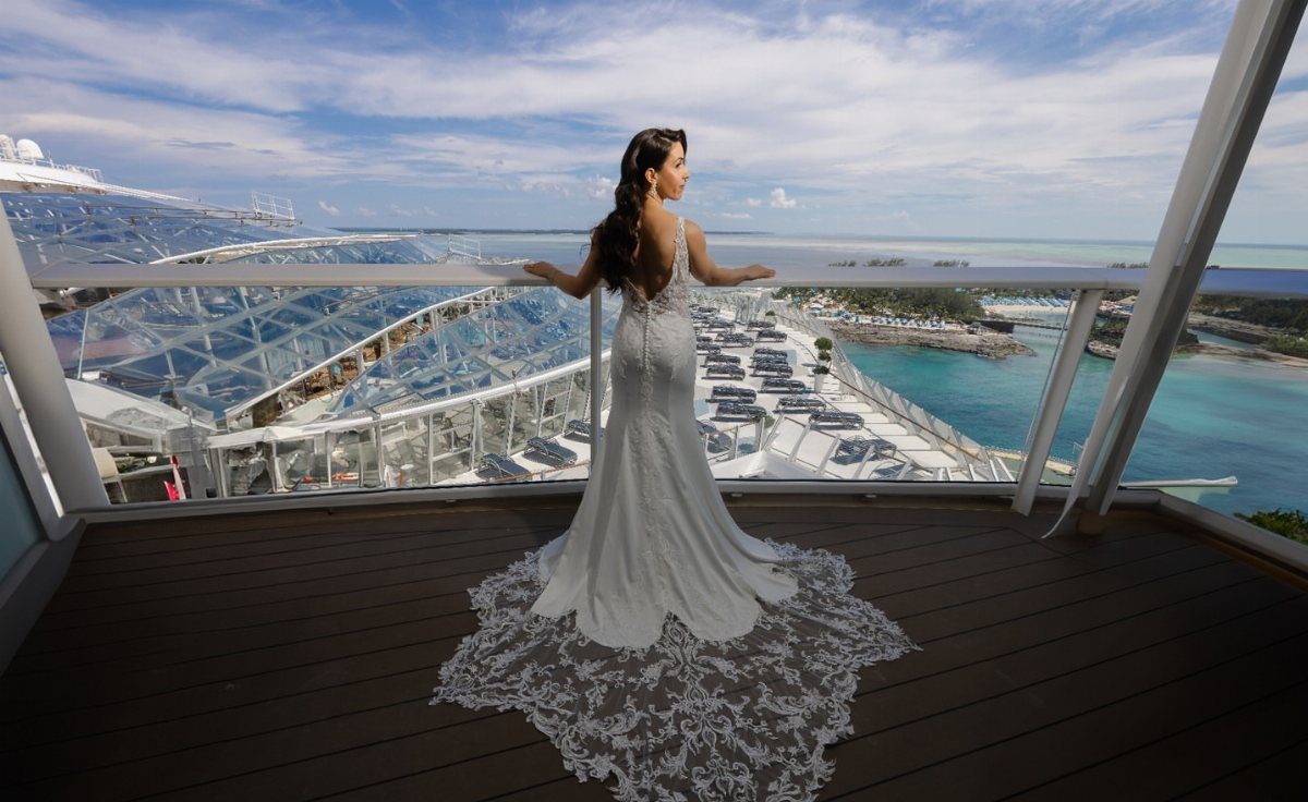 Woman in a white lace wedding dress standing on a balcony overlooking a cruise ship deck.