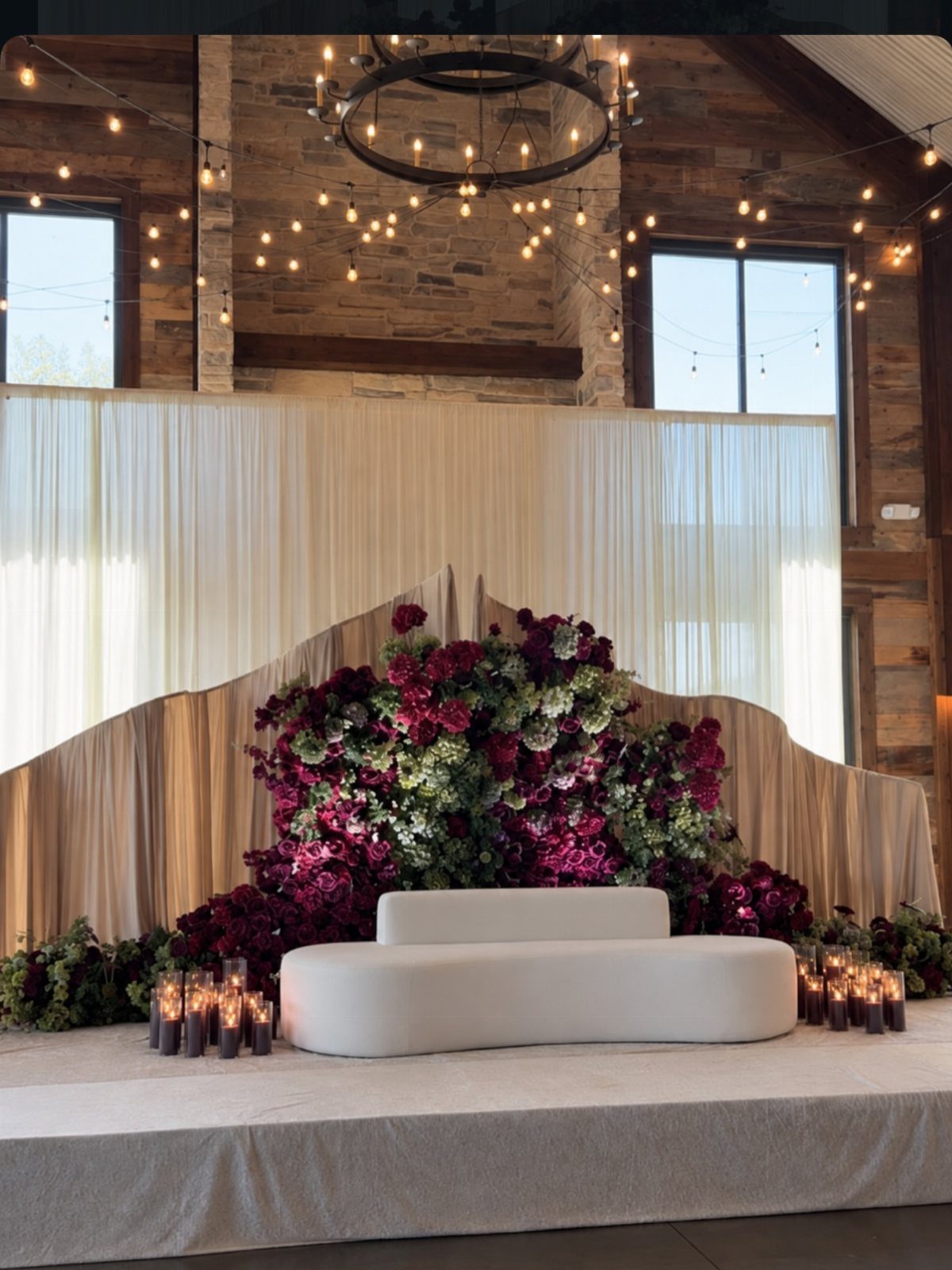 White curved sofa on a platform surrounded by large red and pink floral arrangements.
