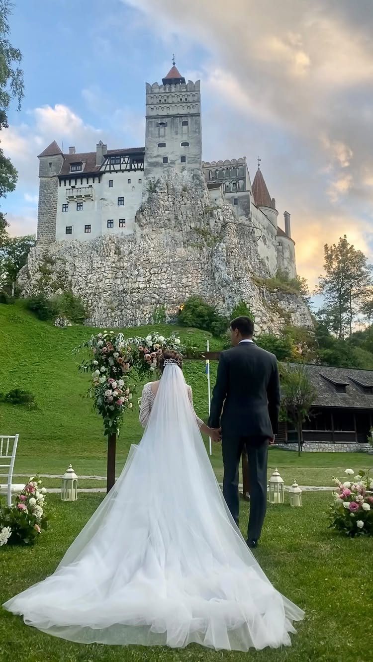 A bride and groom walking toward a floral arch with a large castle in the background.