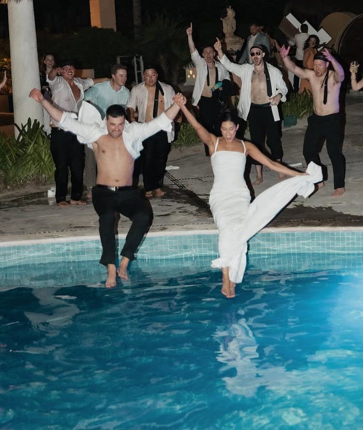 A bride and groom jumping into a swimming pool surrounded by cheering wedding guests at night.