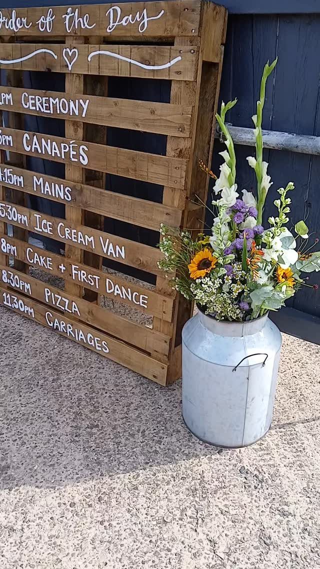 Wildflower Wedding Theme: 13 Ideas for a Light, Natural Day 13 A wooden wedding schedule sign stands next to a milk churn filled with colorful wildflowers.