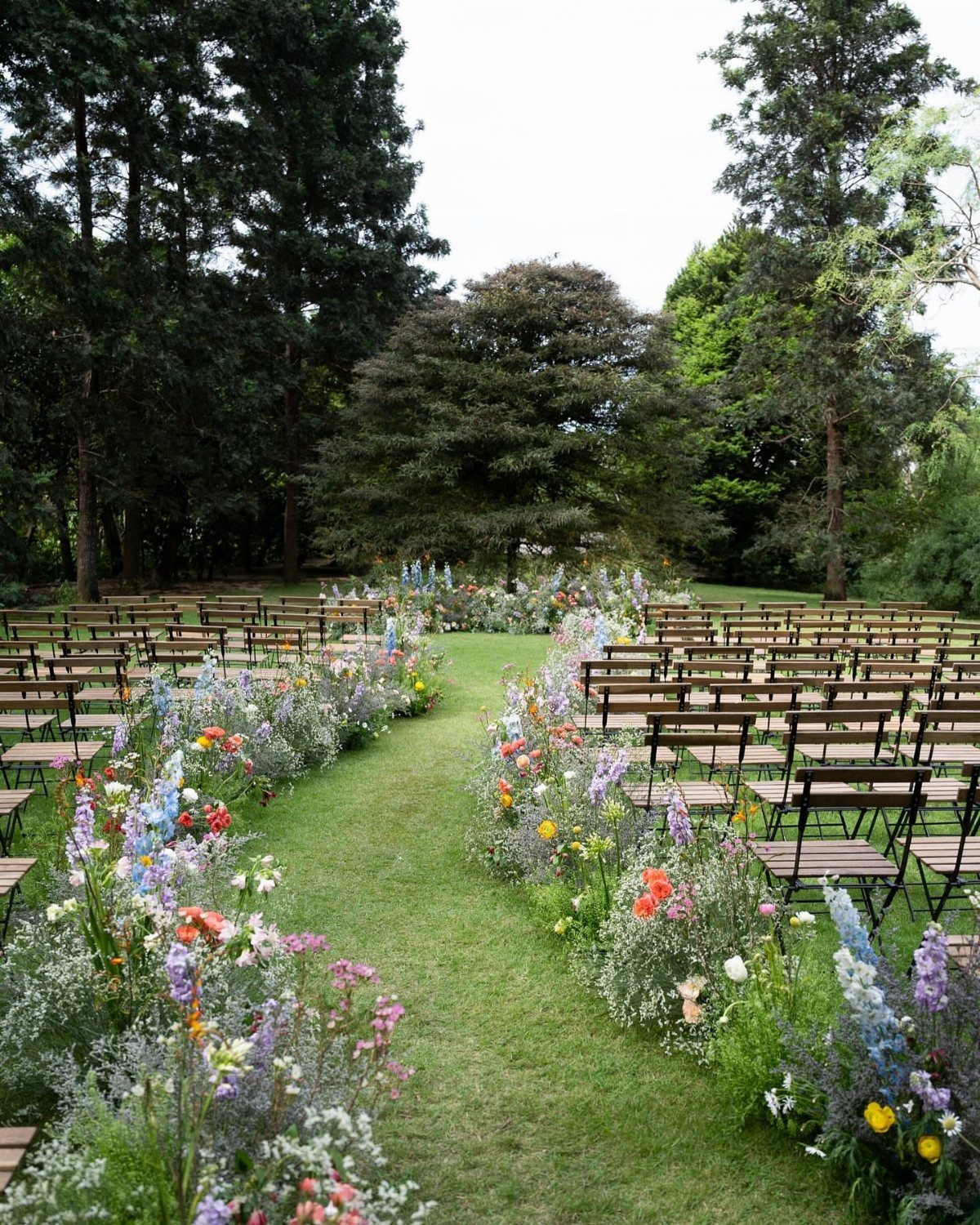 Wildflower Wedding Theme: 13 Ideas for a Light, Natural Day 1 Wildflowers line a grassy wedding aisle flanked by rows of wooden chairs under tall green trees.