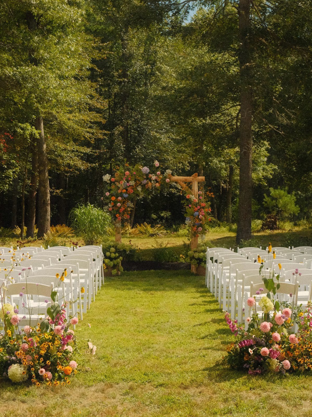 Wildflower Wedding Theme: 13 Ideas for a Light, Natural Day 3 White wedding chairs arranged in rows facing a floral wooden arch in a forest clearing.