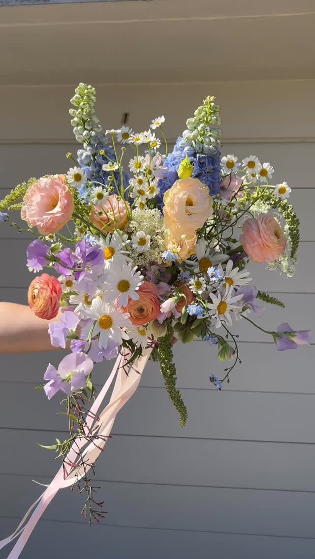 Wildflower Wedding Theme: 13 Ideas for a Light, Natural Day 2 A colorful wildflower bouquet with pink ranunculus, blue delphinium, and white daisies tied with pink ribbons.