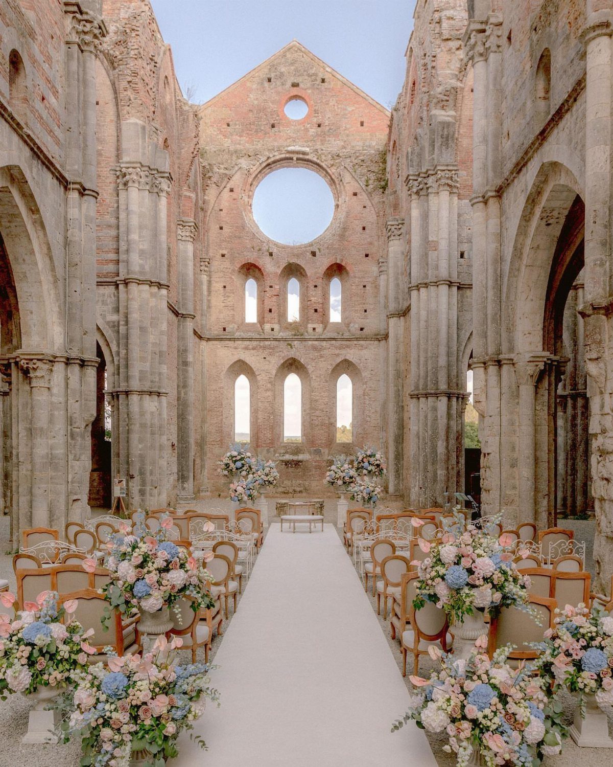 Floral arrangements in dusty blue and blush tones line a white aisle in stone ruins.