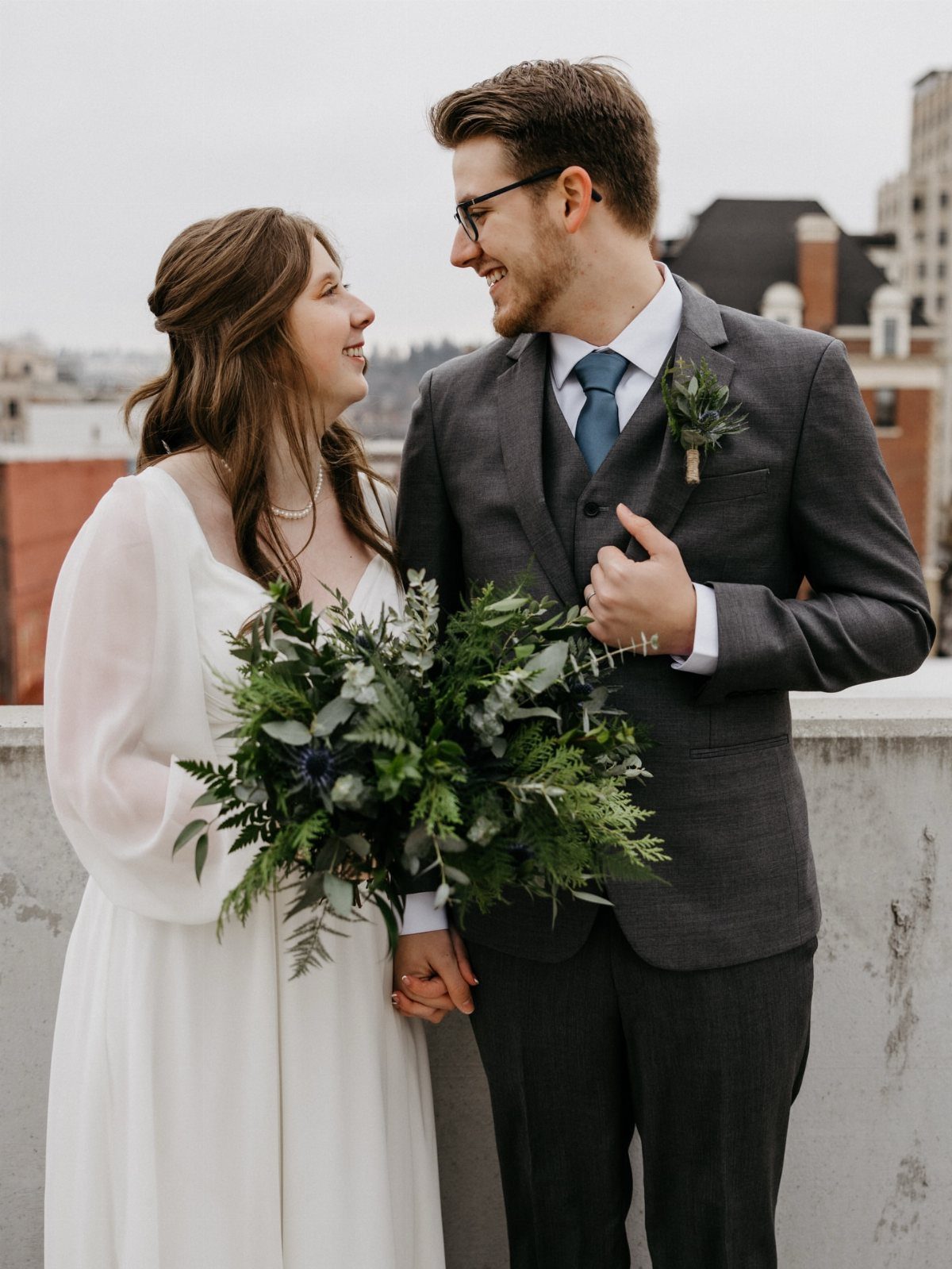 A bride holding a large greenery bouquet and a groom in a grey suit smiling at each other.