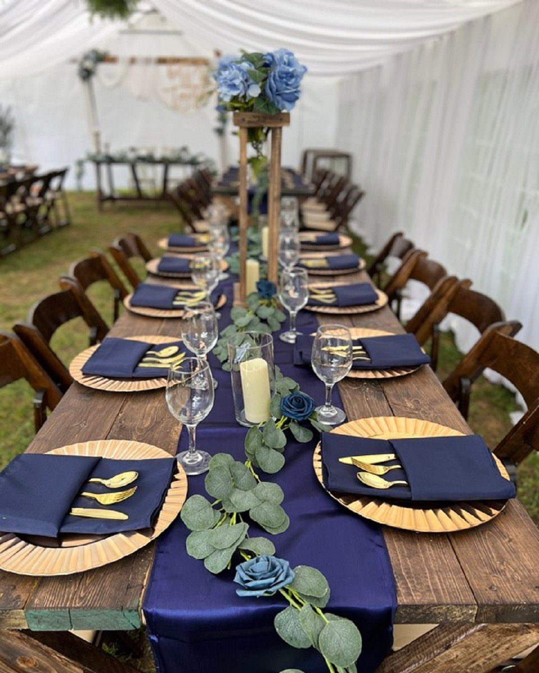 A long wooden wedding table decorated with navy blue runners, gold plates, and green eucalyptus leaves.