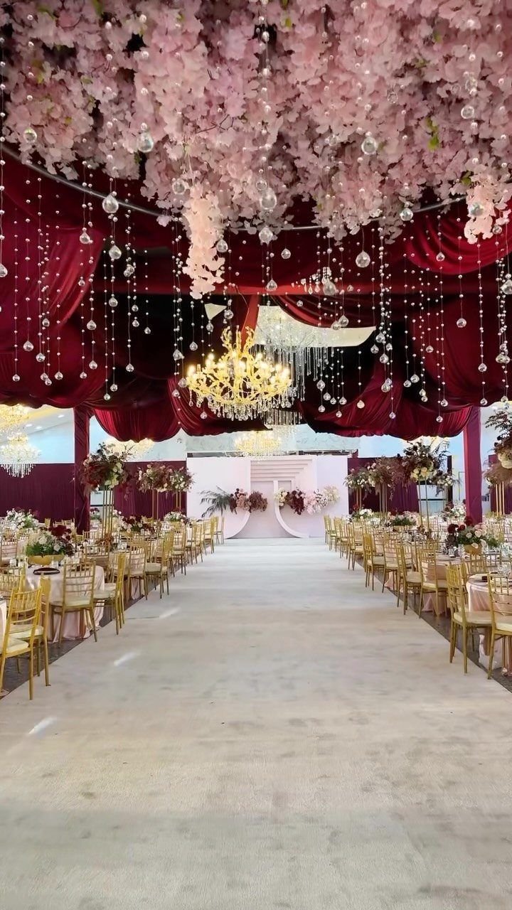 A grand wedding reception hall decorated with red velvet curtains, pink flowers, and gold chairs.