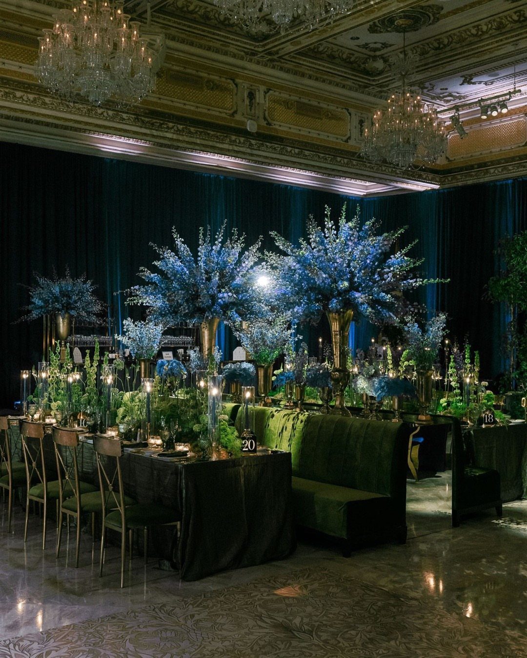 Blue floral centerpieces and green velvet seating in a grand, dimly lit ballroom decorated for a formal event.
