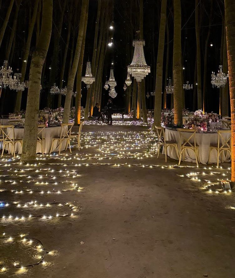 Wedding tables and hanging crystal chandeliers set within a dark forest with lights scattered on the ground.