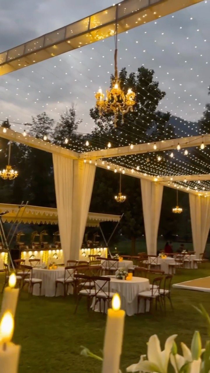 Twinkling fairy lights and golden chandeliers hang over an outdoor wedding reception with white draped tables.