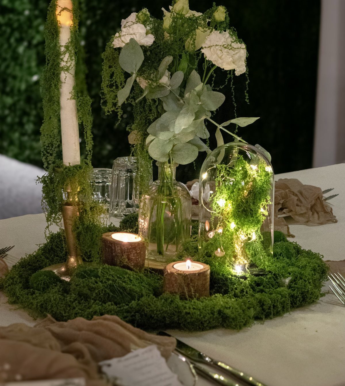 A moss-covered wedding centerpiece with white flowers, candles, and glass vases on a table.