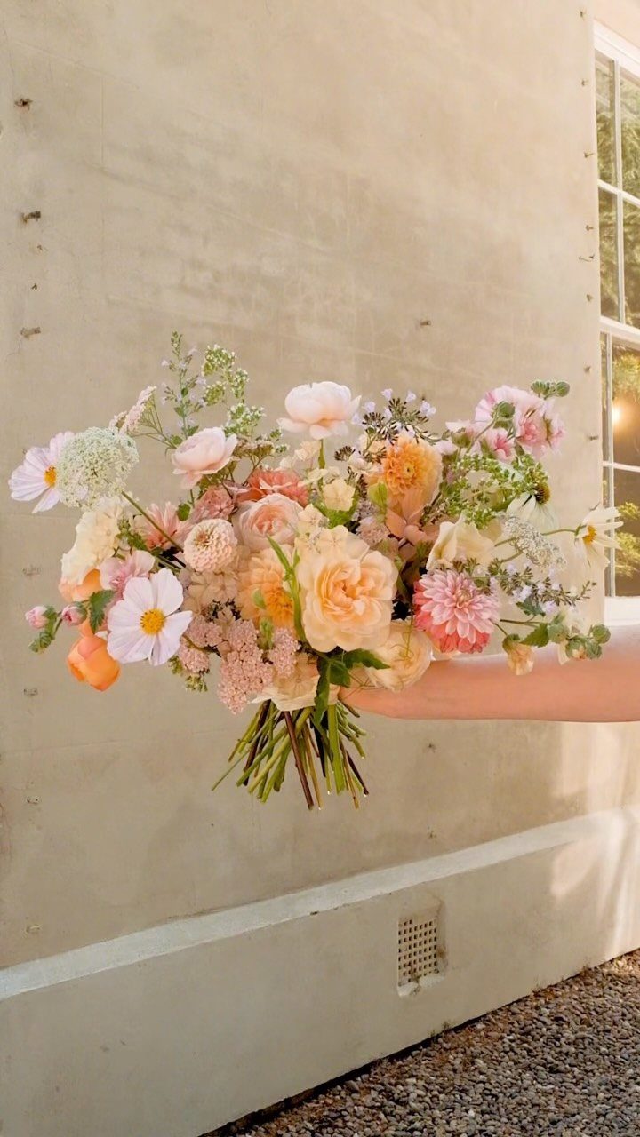 A hand holding a large, colorful bouquet of wildflowers in peach and pink tones.