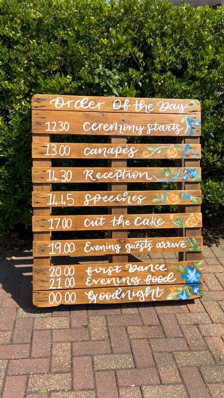 A rustic wooden slat sign displaying a wedding order of the day schedule with floral details.
